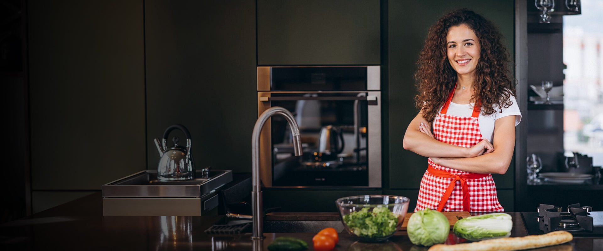 Woman in a kitchen wearing a red apron with various kitchen items and appliances in the background.