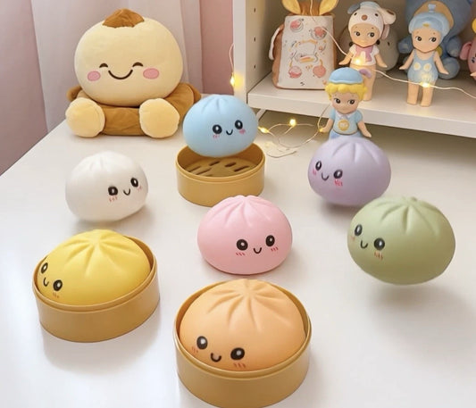 A set of cute, cartoon-faced buns and dumplings in various pastel colors, some displayed inside round bamboo-style steaming baskets and others sitting outside. The set includes yellow, peach, pink, blue, purple, and green items, arranged on a white tabletop with a soft pink background and decorative toys in the distance.